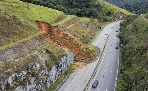 Imagem: Fotografia. Vista do alto de lugar com estrada larga em cinza, passando carros à direita. À esquerda, morro coberto com grama de cor verde-claro e parte com um grande buraco, formando uma cratera onde vê-se o solo marrom. Mais ao fundo, outros morros com vegetação de cor verde. À direita, sobre ruas, carros passam.  Fim da imagem.