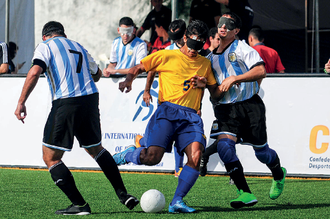 Imagem: Fotografia. Sobre campo de futebol com grama de cor verde, três jogadores de futebol, com os olhos vendados. Dois deles estão com uniforme com camiseta em branco e azul-claro, com bermuda e meiões em preto. À esquerda, um é visto de costas e ao fundo, outros dois são vistos para de frente. Ao centro, um com uniforme do Brasil, camiseta de cor amarela, com bermuda em azul e meiões em branco e chuteiras em azul-claro. Ao fundo, barreira n a horizontal branca e pessoas em pé.  Fim da imagem.