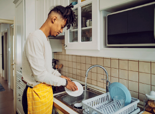 Imagem: Fotografia. Um homem lavando louça de frente para pia, em uma cozinha. Ele tem pele morena, cabelos pretos trançados para cima, usando blusa de mangas compridas em cinza-claro, com um avental amarelo sobre calça preta. Na pia cinza, torneira da mesma cor por onde sai água e à direita, escorredor em cinza-claro com um prato em azul-claro. Na parte superior, armário de cor branca.  Fim da imagem.