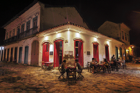 Imagem: Fotografia. No céu noturno em preto, com casarões de paredes brancas, com portas em vermelho. À esquerda e à direita, com dois andares, com janelas de cor azul. No teto, luz branca. De frente para as portas, mesas pequenas com cadeiras e pessoas comendo.  Fim da imagem.
