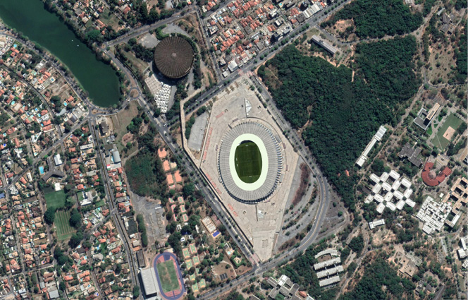 Imagem: Fotografia aérea. Ao centro, um local arredondado cinza, com parte verde dentro dele e ao redor, local cinza. No entorno, casas, prédios e ruas longas de cor cinza. Na parte esquerda, rio de água de cor verde e à direita, árvores com folhas verdes.  Fim da imagem.