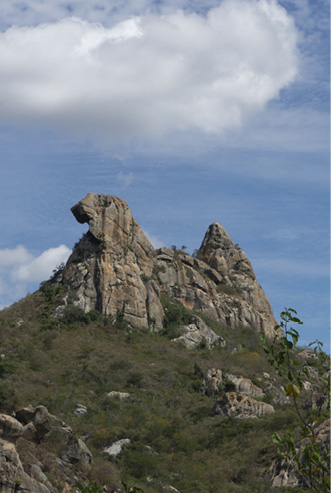 Imagem: Fotografia. Vista geral de local com uma pedra gigante de cor cinza no alto, sobre morro com vegetação verde. Na parte superior, céu de cor azul com nuvens brancas.  Fim da imagem.