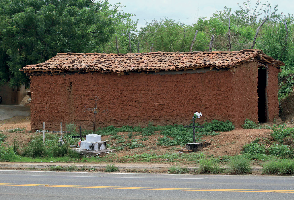 Imagem: Fotografia. À frente, rua de cor cinza com linhas finas em amarelo na horizontal. Em segundo plano, solo de cor marrom, vegetação verde, uma casa grande de cor marrom-claro, com porta grande na frente, e no alto, telhado triangular telhas em marrom. Mais ao fundo, atrás da casa, árvores verdes. Na ponta superior, em azul-claro.  Fim da imagem.