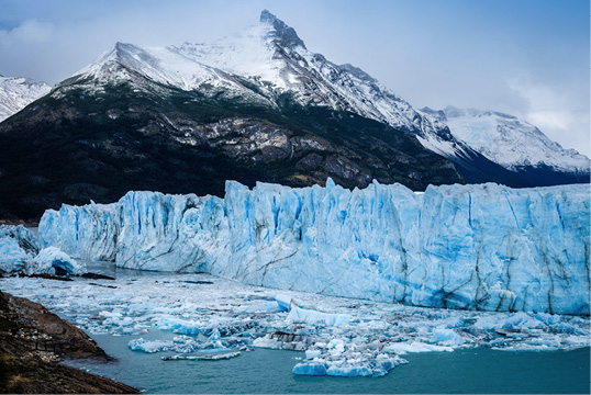 Imagem: Fotografia de grandes paredes de gelo. Em frente, lago com blocos de gelo. Ao fundo, montanha com neve no topo. Fim da imagem.