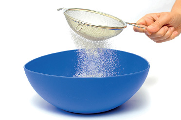 Imagem: Fotografia. Passo 1. Mão de uma pessoa de pele clara, com peneira de cor cinza, peneirando com farinha branca sobre uma tigela de cor azul.  Fim da imagem.