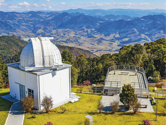 Imagem: Fotografia. Na parte inferior, local com grama de cor verde-claro. À esquerda, um local com paredes em branco, com a parte superior, uma cúpula arredondada da mesma cor. À direita, local com árvore de folhas verdes, grades finas na horizontal e ao fundo, local aberto com grade contornando em cinza. Em segundo plano, muitas árvores com folhas verdes e mais ao fundo, morros de tamanhos diferentes, em marrom e verde. Na parte superior, céu azul-claro com nuvens brancas.  Fim da imagem.