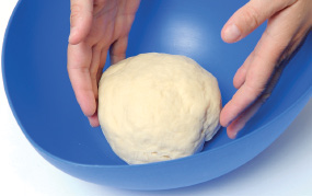 Imagem: Fotografia. Passo 5. Mãos de uma pessoa colocando a massa com o formato arredondado de cor bege-claro.  Fim da imagem.