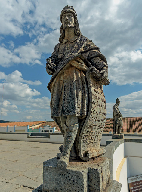 Imagem: Fotografia. Escultura em cinza, sobre uma pedra, de homem na vertical, com barba, nariz fino e queixo mais quadrados, com vestes antigas, com casaco até os joelhos, botas longas e à direita, segura na mão direita um pergaminho com texto. Ao fundo, outra escultura de pessoa em pé sobre pedra. Na parte superior, céu azul-claro com nuvens brancas.  Fim da imagem.