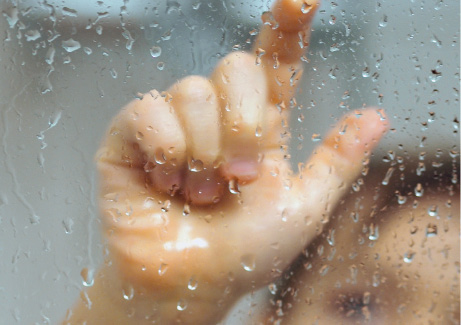 Imagem: Fotografia. Sobre vidro transparente com gotículas de água, com a mão esquerda para frente, com dedo indicador com polegar para cima e cabeça à direita, vista parcial de cabeça de pele clara, com cabelos castanhos.  Fim da imagem.