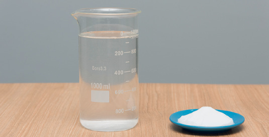 Imagem: Fotografia. Sobre uma base de madeira em bege, um frasco transparente arredondado com água dentro. À direita, base arredondada em azul com pó branco. Mais ao fundo, parede de cor cinza.  Fim da imagem.