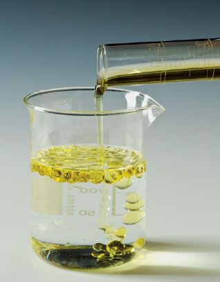 Imagem: Fotografia. À esquerda, um frasco transparente arredondado, com água com frasco em cima transparente, na horizontal, óleo de cor amarela, fazendo folhas na água.  Fim da imagem.