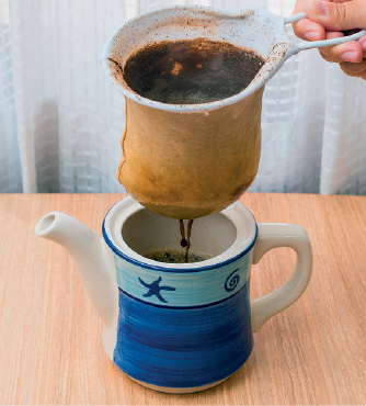 Imagem: Fotografia. Mão de uma pessoa de pele clara, segurando um filtro branco, com pó dentro de cor marrom, escorrendo café dentro de jarra de cerâmica branca, com detalhes em azul-escuro e parte em azul-claro, sobre uma base de madeira.  Fim da imagem.