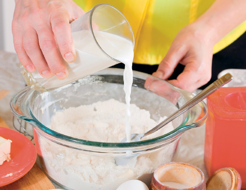Imagem: Fotografia. Mãos de uma pessoa de pela clara, com roupa amarela, segurando copo transparente na mão esquerda com leite, despejando-o sobre massa branca dentro de vasilha de vidro transparente, com colher à direita.  Fim da imagem.