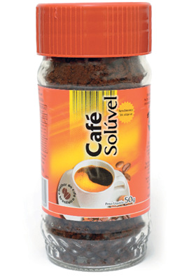 Imagem: Fotografia. Um pote de vidro transparente, com pó de cor marrom-escuro, com etiqueta de cor laranja sobre o pote, com xícara com pires em branco. Na parte superior, tampa laranja arredondada.  Fim da imagem.