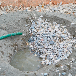 Imagem: Fotografia. Cimento de cor cinza-claro, com pedras de cor branco e cinza e mangueira à esquerda de cor verde por onde sai água.  Fim da imagem.
