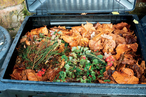 Imagem: Fotografia. Vista do alto de caixa preta com a tampa para cima em cinza. Dentro da caixa, restos de comida e vegetais com folhas verdes.  Fim da imagem.