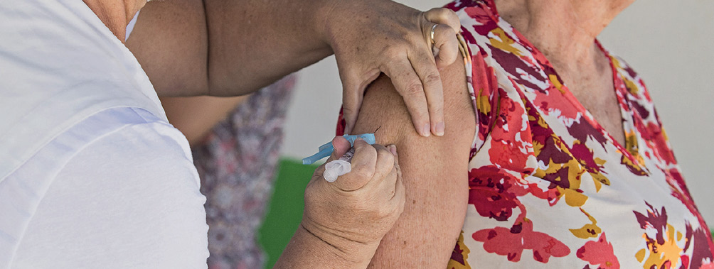 Imagem: Fotografia. À esquerda, vista parcial de uma pessoa de pele clara, com roupa de cor branca, com a mão esquerda sobre o ombro de uma mulher e mão direita com seringa sobre braço de mulher. A mulher tem pele clara, com blusa regata em branco, com partes floridas em vermelho e amarelo.  Fim da imagem.