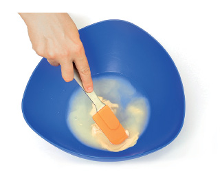 Imagem: Fotografia. Passo 2. Vista do alto para cima, mão de uma pessoa de pele clara, à esquerda, segurando uma espátula mistura de água com ingredientes.  Fim da imagem.