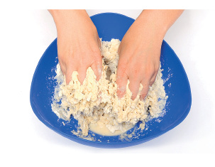 Imagem: Fotografia. Passo 3. Mãos de uma pessoa de pele clara, amassando massa de cor bege-claro com textura pegajosa. Fim da imagem.