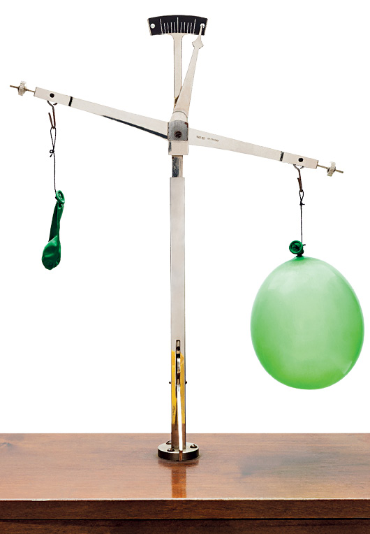 Imagem: Fotografia. Sobre base na horizontal de cor marrom-escuro. Sobre a base, objeto com haste fina branca na vertical com ponta inferior em dourado e haste fina da mesma cor na horizontal. À esquerda, com a ponta para cima, com bexiga murcha em verde. Na ponta da direita, bexiga de cor verde, arredondada.  Fim da imagem.