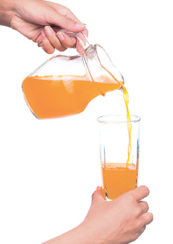 Imagem: Fotografia. Mãos de uma pessoa de pele clara, segurando na mão esquerda, jarra de cor transparente com suco laranja, colocando em copo transparente segurado na mão direita.  Fim da imagem.