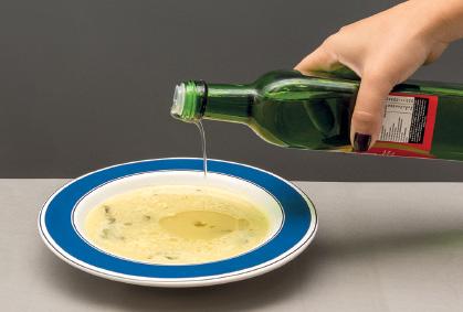 Imagem: Fotografia. Mão de uma pessoa de pele clara, de unhas com esmalte preto, segurando uma garrafa de cor verde, jorrando azeite sobre sopa de cor bege-claro, em um prato redondo com contorno em azul-escuro.  Fim da imagem.