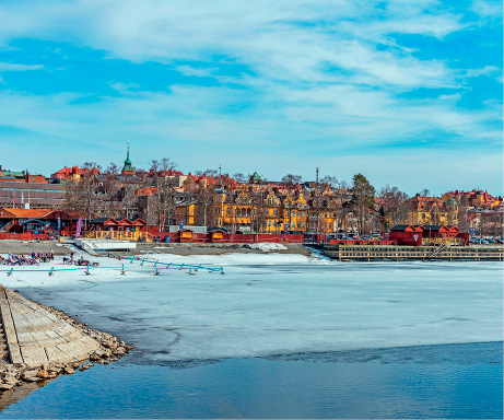 Imagem: Fotografia. À frente, rio de cor azul-claro, grande e mais ao fundo, cidade com prédios pertos um dos outros. Na parte superior, céu de cor azul-claro com nuvens brancas esparsas.  Fim da imagem.