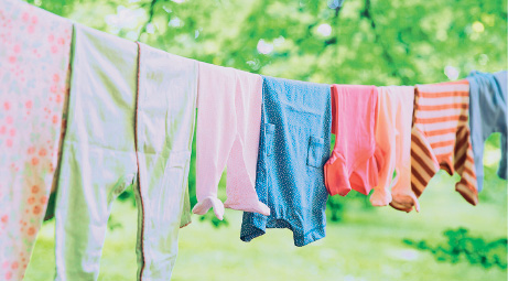 Imagem: Fotografia. Um varal com roupas estendidas em verde, bege, azul, vermelho, laranja e bege com marrom. Mais ao fundo, vegetação de cor verde.  Fim da imagem.