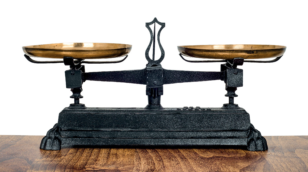 Imagem: Fotografia. Balança antiga, na horizontal de cor preta, com duas hastes em cada ponta. À esquerda e à direita, pratos arredondados dourados. Ao centro, haste na vertical preta.  Fim da imagem.