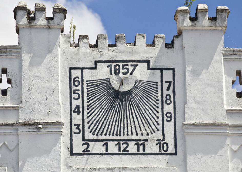 Imagem: Fotografia. Vista da frente de uma fortaleza de cor cinza-claro, com relógio com listras em preto ao centro. Ele tem forma quadrada, com data na parte superior : 1857. Ao redor, linhas finas na vertical e numeração em sentido anti-horário: 6, 5, 4, 3, 2, 1, 12, 11, 10, 9, 8, 7. Na parte superior, céu azul-claro com nuvem à esquerda em branco.  Fim da imagem.