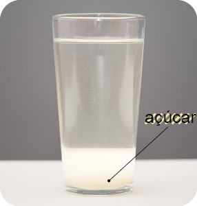 Imagem: Fotografia. Um copo transparente arredondado com água. Na parte inferior, açúcar com parte da água em branco no fundo do copo.  Fim da imagem.