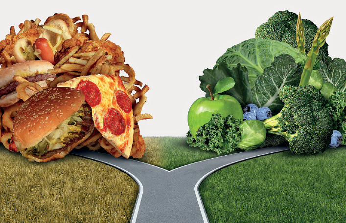 Imagem: Fotografia. Ao centro, rua de cor cinza, com bifurcação à frente e à esquerda e à direita. À esquerda, grama seca e na ponta, visto em escala grande, um amontoado de lanches:  sanduíches, pedaço de pizza de queijo, batatas-fritas; cebolas fritas e salsicha. À direita, grama de cor verde e na ponta, amontoado de vegetais, brócolis, uma maçã de cor verde, visto em escala grande.  Fim da imagem.