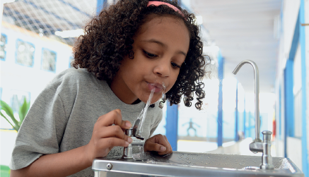 Imagem: Fotografia. Uma menina vista da cintura para cima, de pele morena, com cabelos curtos encaracolados em castanho, com camiseta de cor cinza. Ela está de frente para bebedouro de cor cinza, com pequena saída na vertical por onde jorra água perto da boca da menina. À direita, ao fundo, torneira fina curvada para baixo.  Fim da imagem.