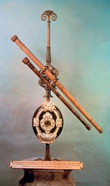 Imagem: Fotografia. Na vertical, de cor marrom, com luneta na diagonal de cor marrom, com parte menor e outra maior de formato tubular. Na vertical, detalhes ornamentados antigos. Fundo de parede de cor azul-claro.  Fim da imagem.