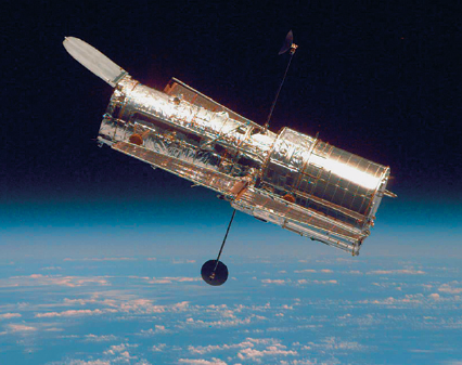 Imagem: Fotografia. No alto, acima de céu azul-claro e com nuvens brancas, um telescópio na diagonal, forma tubular de tamanho médio, com a ponta à esquerda, com haste fina na vertical em preta.  Fim da imagem.