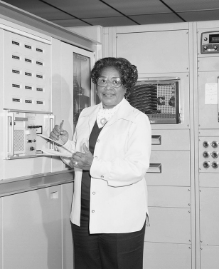 Imagem: Fotografia. Uma mulher em pé vista dos joelhos para cima, de pele escura, cabelos escuros, com blusa de mangas compridas em branco e saia escura. Ela usa um par de óculos no rosto, com prancheta nas mãos anotando. Ao fundo, local com paredes claras.  Fim da imagem.