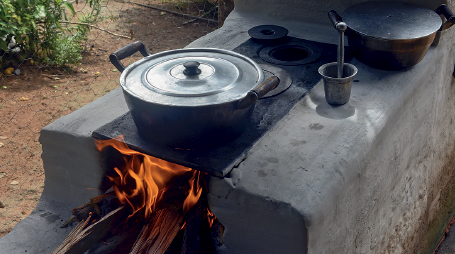 Imagem: Fotografia. Vista do alto de um fogão à lenha, estrutura feita com base na horizontal em cinza, abertura em baixo por vê-se chamas de cor laranja. Na parte superior, base na horizontal em preto e uma panela redonda em cinza-escuro com tampa. Próximo, à direita, um pilão, utensílio semelhante a copo e objeto que amassa temperos. Ao fundo, à esquerda, local com solo marrom e vegetação verde.  Fim da imagem.