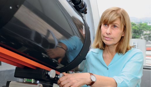 Imagem: Fotografia. Uma mulher vista do busto para cima, cabelos curtos longos, com franja na frente, com blusa de mangas até os cotovelos de cor azul-claro. Ela olha para frente e segura com a mão direita sobre um telescópio grande de cor preta. Ao fundo, vista parcial de cidade. Fim da imagem.