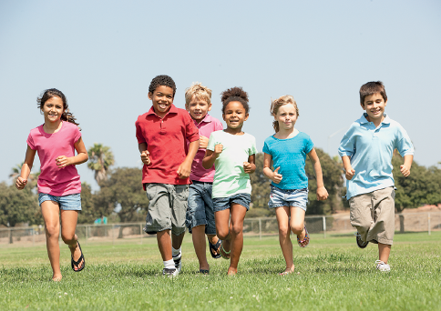 Imagem: Fotografia. Em um local aberto com grama de cor verde, seis crianças correndo para frente. Uma menina de pele clara, cabelos presos de cor castanha, com camiseta rosa, short azul-claro e par de chinelos pretos. Mais à direita, um menino de pele negra, cabelos pretos, com camiseta vermelha, bermuda cinza; um menino loiro com camiseta rosa, bermuda cinza; uma menina negra, com cabelos castanhos crespos, com camiseta de cor cinza-claro, short azul; uma menina loira, camiseta azul, com short cinza e na ponta direita, um menino de pele clara, cabelos castanhos, com camiseta azul-claro e bermuda longa em cinza. Em segundo plano, árvores com folhas verdes. Na parte superior, céu azul-claro sem nuvens.  Fim da imagem.