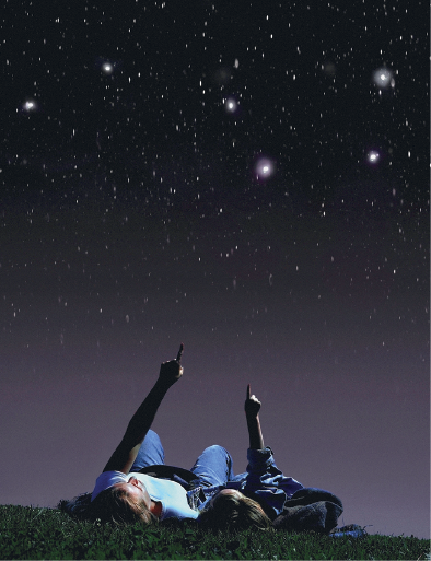 Imagem: Fotografia. Sobre local com grama verde, uma mulher à esquerda, com blusa branca, calça jeans, com o dedo indicador da mão esquerda apontando para o alto. À direita dela, uma menina deitada com o dedo indicador da mão direita para cima. No céu preto, com parte inferior em cinza. Na parte preta, estrelas, centenas de esferas brancas, pequenas, médias e algumas grandes.  Fim da imagem.