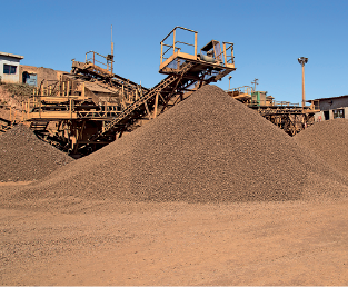 Imagem: Fotografia. Vista local com três montes de cor marrom, com formato triangular. Ao fundo, um trator de ferros de cor amarela. Mais ao fundo, outras partes em ferro.  Fim da imagem.