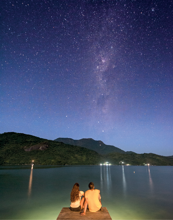 Imagem: Fotografia. Na parte inferior, uma mulher de cabelos longos castanhos, blusa branca e bermuda preta e um homem de pele clara, cabelos castanhos, com camiseta bege, sentados em base de madeira, vistos de costas. Eles estão de frente para local com água. Em segundo plano, morros cobertos de cor verde. Na parte superior, céu azul com centenas de estrelas brancas, pequenas.  Fim da imagem.