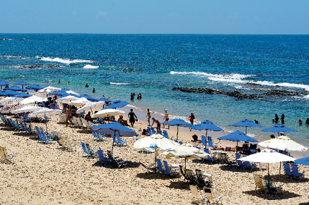 Imagem: Fotografia. Praia de cor bege-claro, com guarda-sol em azul e branco, com cadeiras. À frente, pessoas sentadas e outras caminhando. Mais ao fundo, mar de água de cor azul com ondas. Algumas pessoas estão na borda da praia.  Fim da imagem.