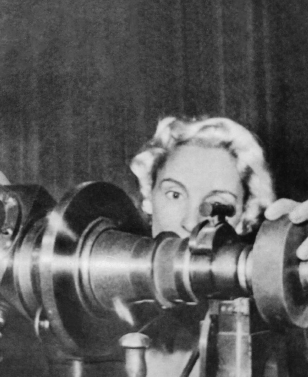 Imagem: Fotografia. Atrás de um objeto na horizontal escuro, com mulher vista parcialmente, com rosto com cabelos curtos claros. Ela tem pele clara e sobrancelhas finas escuras, curvadas, com a mão direita sobre objeto à frente.  Fim da imagem.