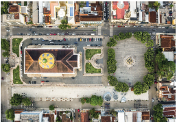 Imagem: Fotografia. Vista de cima de local com ruas de cor cinza, casas na parte de cima e de baixo e ao centro, vista do alto de teatro com telhado marrom e cúpula de cor amarela. À direita, local com árvores de folhas verdes.  Fim da imagem.