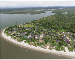 Imagem: Fotografia. Vista geral do alto de local com mar escuro à esquerda e à direita, com duas embarcações, local com solo de cor bege-claro, árvores de folhas verdes, casas e construções baixas. Em segundo plano, densa vegetação e em segundo plano, mais territórios com outras vegetações. No alto, céu com nuvens esparsas em cinza-claro. Fim da imagem.