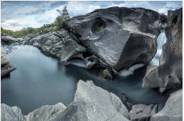 Imagem: Fotografia. Rochas de cor cinza, à direita, uma queda água caindo para lugar com água parada de cor escura. À esquerda, em segundo plano, local com vegetação verde. No alto, céu azul com muitas nuvens. Fim da imagem.