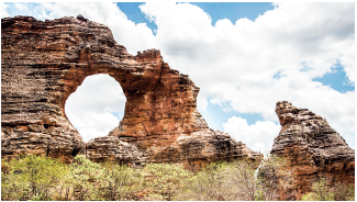 Imagem: Fotografia. Uma rocha marrom no alto, à esquerda, com o buraco arredondado ao centro. À direita, outra pequena rocha da mesma cor na vertical. No alto, céu azul e nuvens brancas. Fim da imagem.
