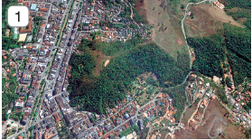 Imagem: Fotografia 1. Vista do alto de local com ruas e prédios e casas à esquerda. À direita, local som solo marrom, grande parte com vegetação e árvores de folhas em verde-escuro. Fim da imagem.