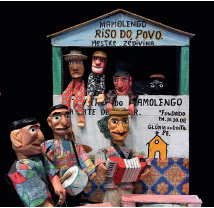 Imagem: Fotografia. À frente, três bonecos de cor marrom, com cabelos escuros, roupas coloridas, com instrumentos musicais nas mãos: um tambor pequeno branco, outro tocando sanfona vermelha. Em segundo plano, casa de paredes em verde, forma triangular acima em azul-claro e telhado marrom. Texto: Mamolengo Riso do povo mestre Zédivina. Na janela da casa, outros quatro bonecos com braços compridos: um de pele negra, com chapéu bege e blusa em vermelho. Ao lado, um homem de pele clara, com blusa em cinza e preto; outro de pele morena, cabelos pretos e blusa de mangas compridas em marrom e à direita, rosto de pessoa vista parcialmente de pele negra, olhos brancos com íris preta e sobrancelhas brancas. Ao fundo, parede em preto.  Fim da imagem.